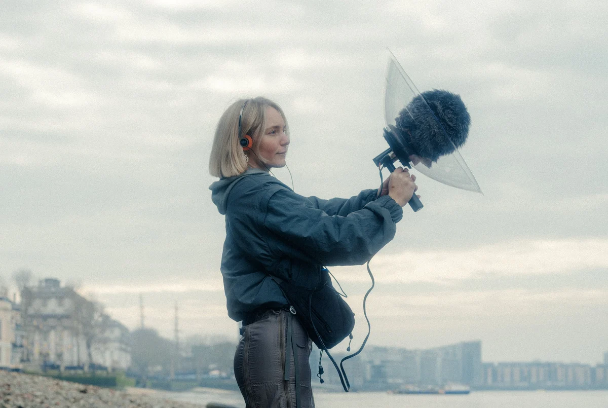 Laura Misch wearing headphones and holding a parabolic microphone on a hazy shoreline